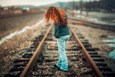 一位橙色头发的女士，穿着黑色和灰色条纹长袖衬衫和浅蓝色牛仔裤，白天站在火车轨道上